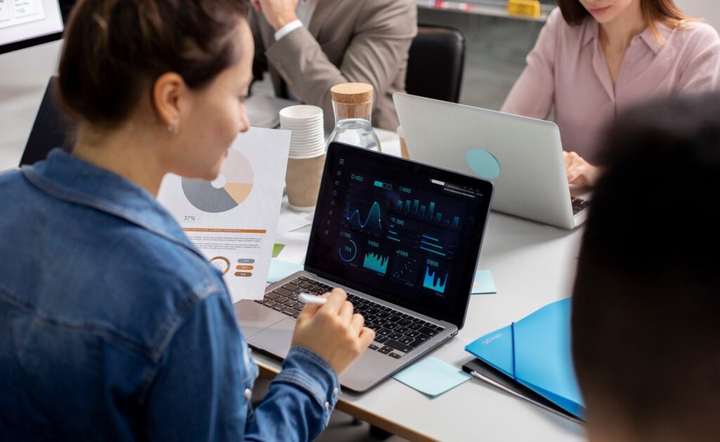 A person in a denim jacket analyzes data on a laptop, surrounded by charts, coffee cups, and colleagues during a meeting.