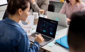 A person in a denim jacket analyzes data on a laptop, surrounded by charts, coffee cups, and colleagues during a meeting.
