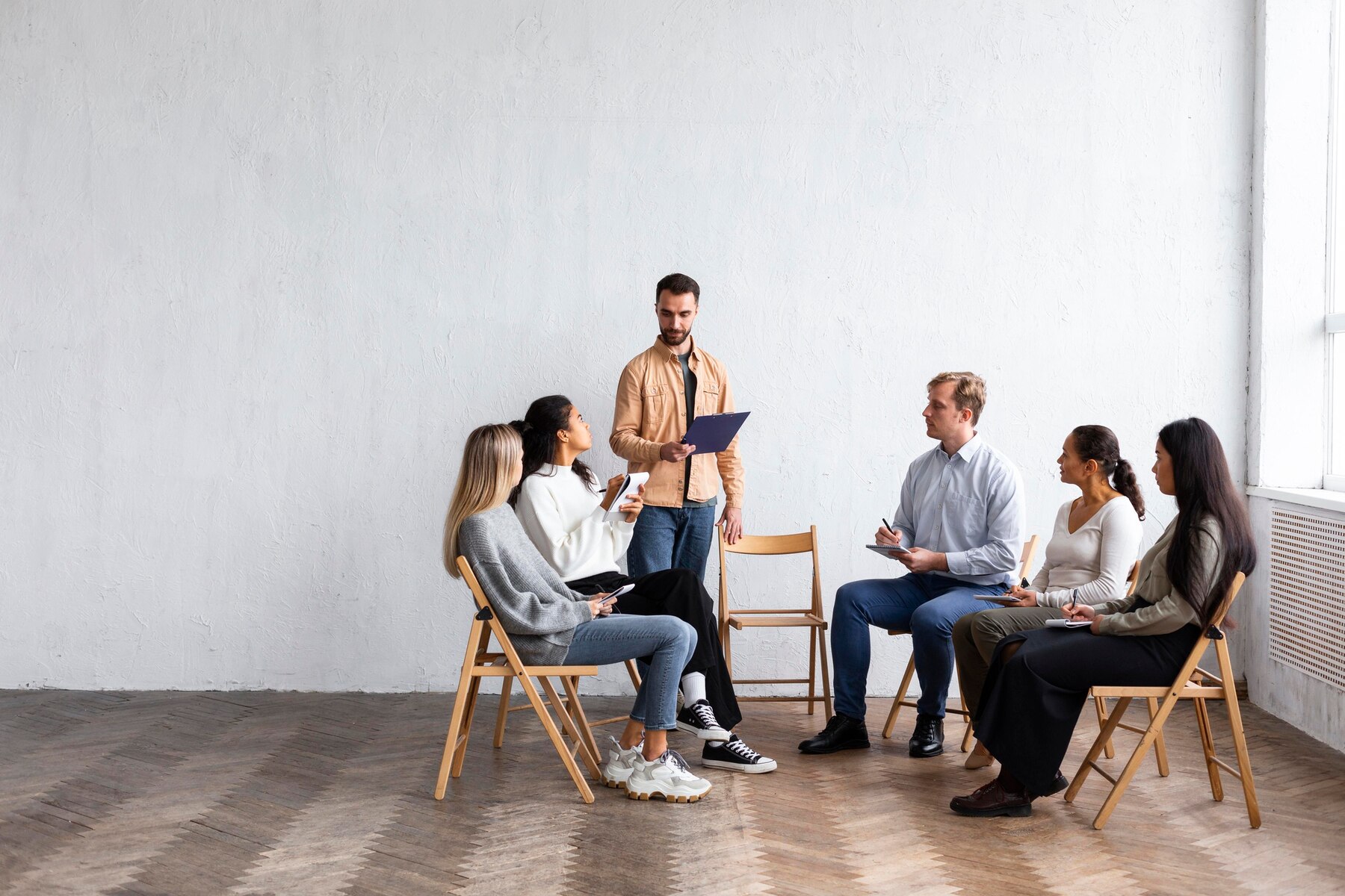 A group of six people seated in a bright room, engaged in a discussion, with one person presenting a document.