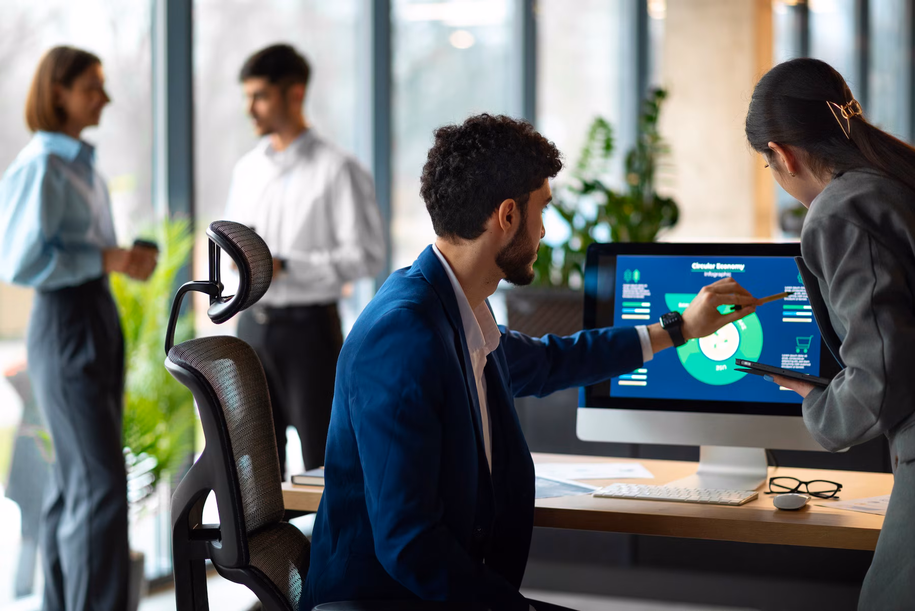 A professional workspace where a man in a suit interacts with data on a screen, while colleagues discuss in the background.