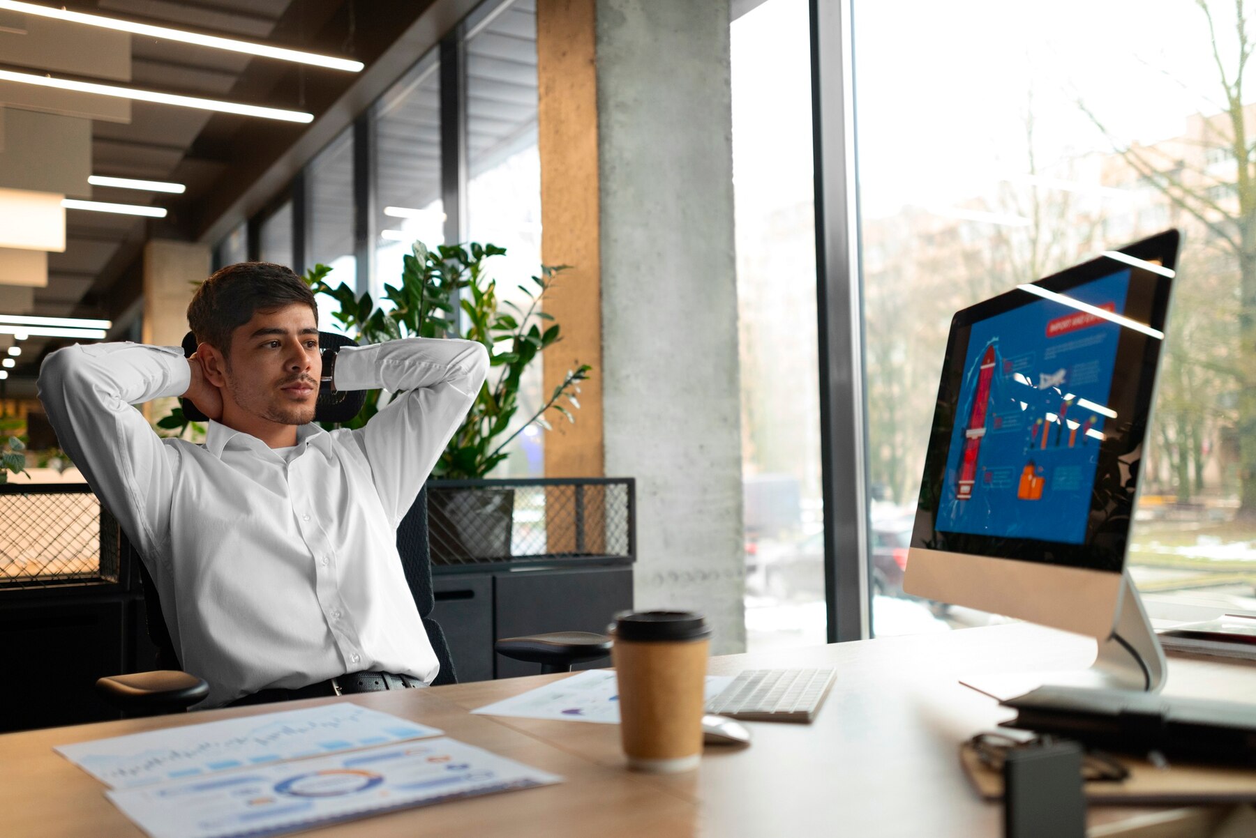 A person in a white shirt leans back in an office chair, looking at a computer screen with digital content, surrounded by greenery.