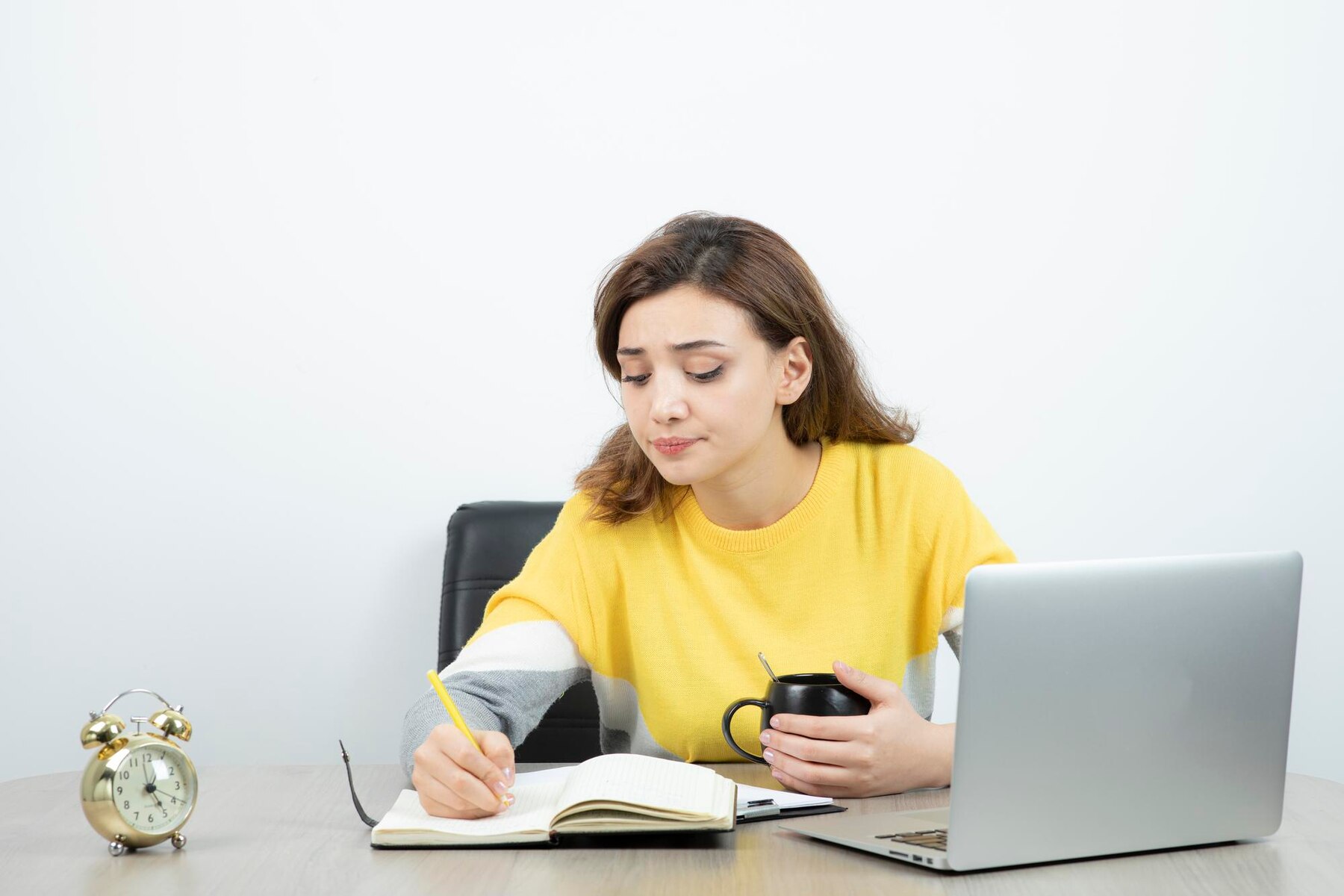 A young woman in a yellow sweater sits at a desk, writing in a notebook and holding a cup, with a laptop and an alarm clock beside her on the table.