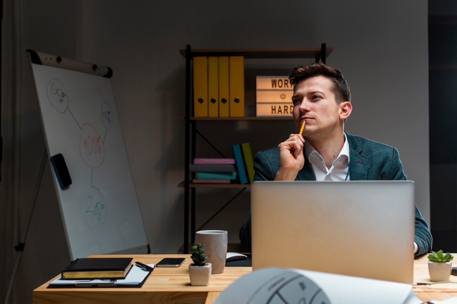 A man sits thoughtfully at a wooden desk with a laptop, surrounded by office supplies, plants, and a whiteboard, in a modern workspace.