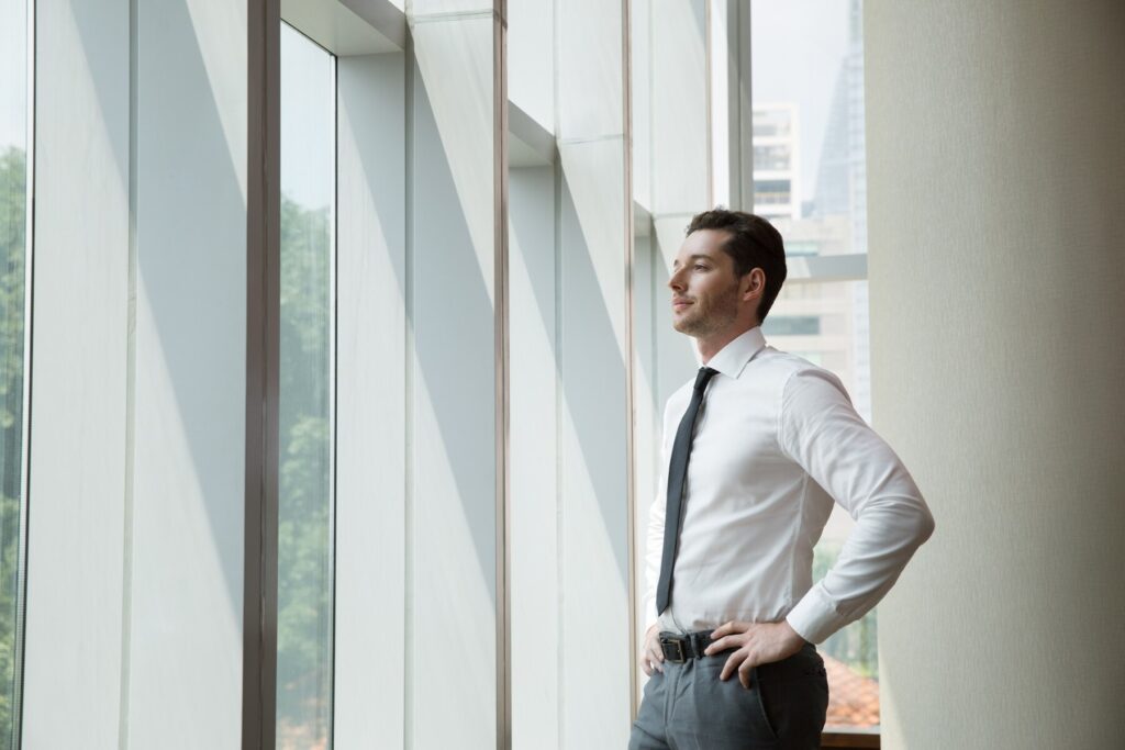 A confident young man in a white shirt and tie stands by large windows in an office, looking contemplatively outside. Natural light illuminates the spacious room.