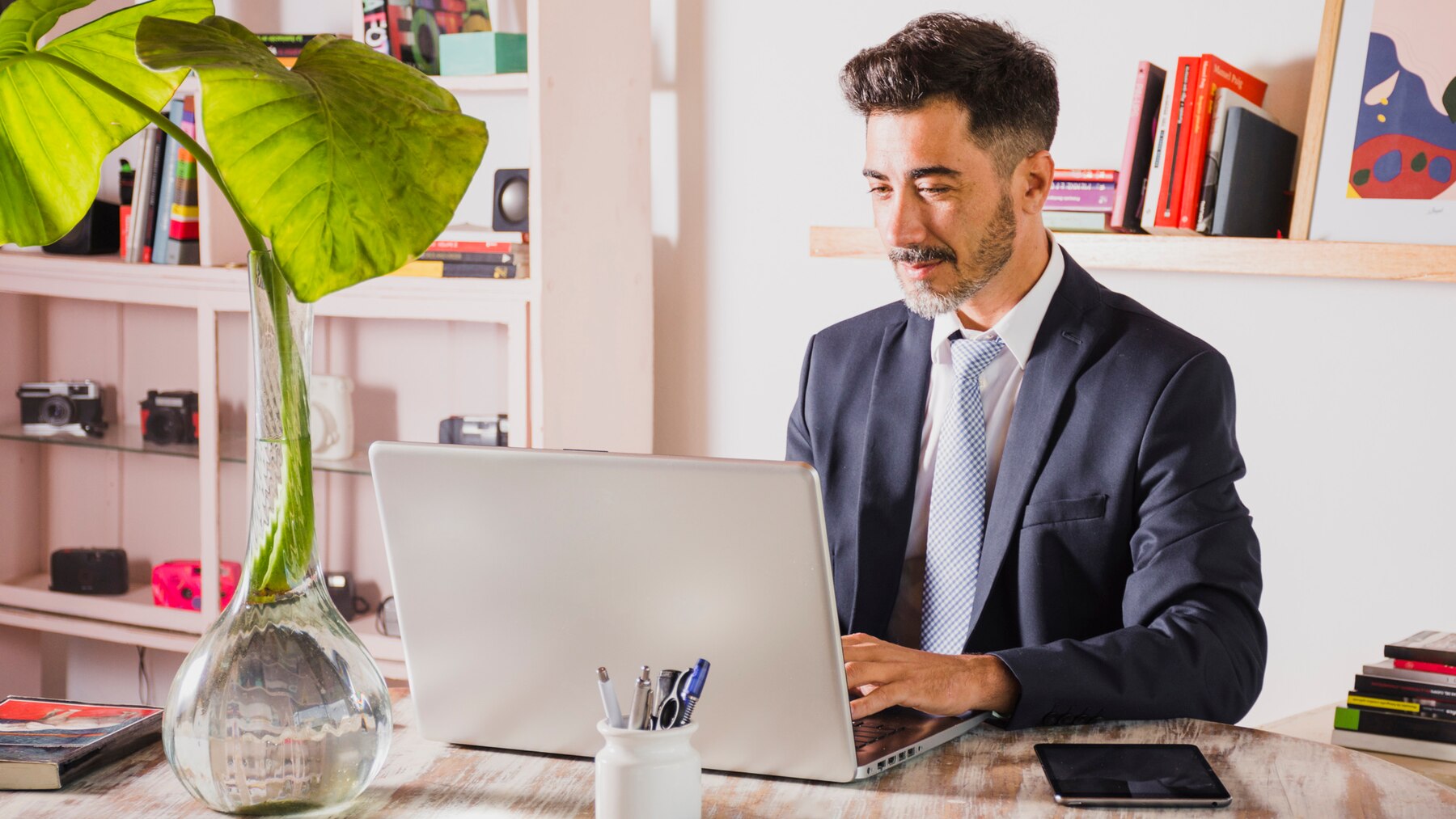 A man in a suit working at a laptop in a stylish home office with a large leafy plant and a shelf filled with books and cameras in the background.