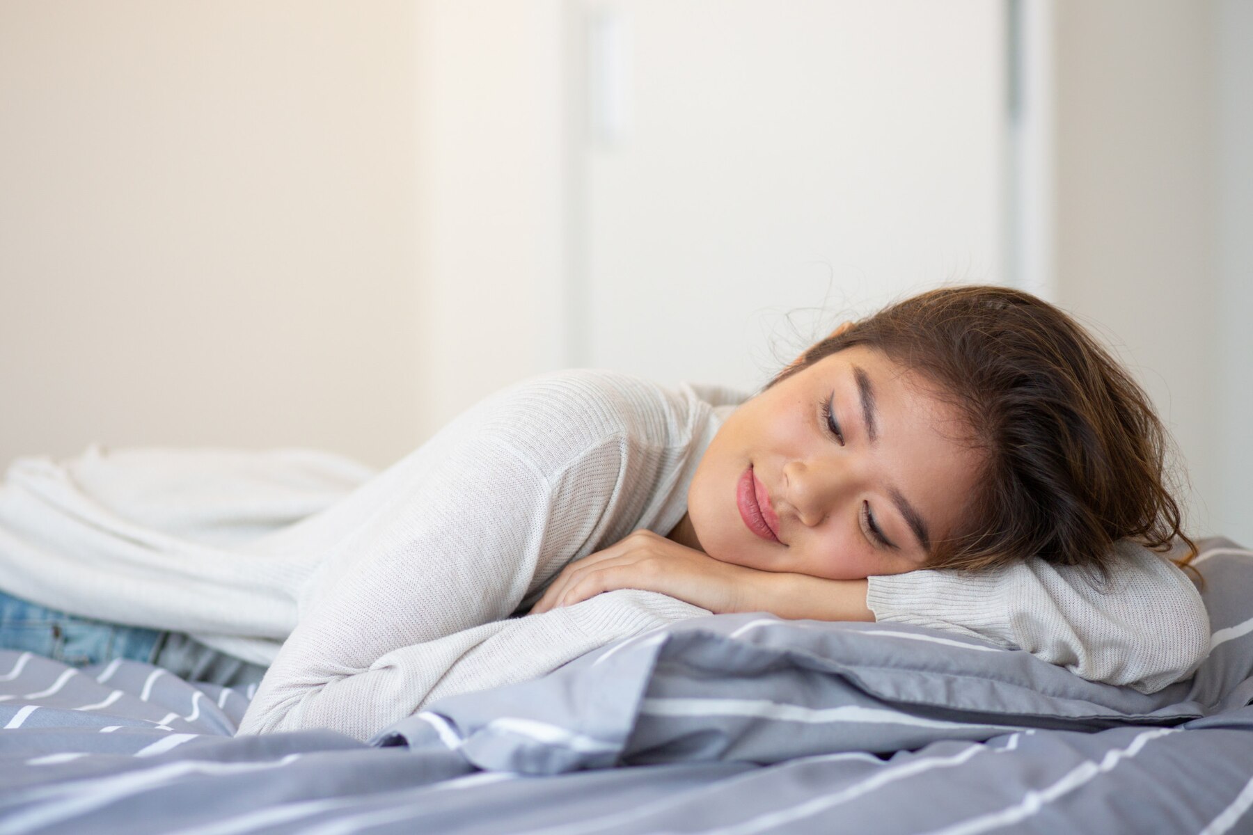 A young woman peacefully sleeping on a bed with light grey and white striped bedding, wearing a comfortable long sleeve top.