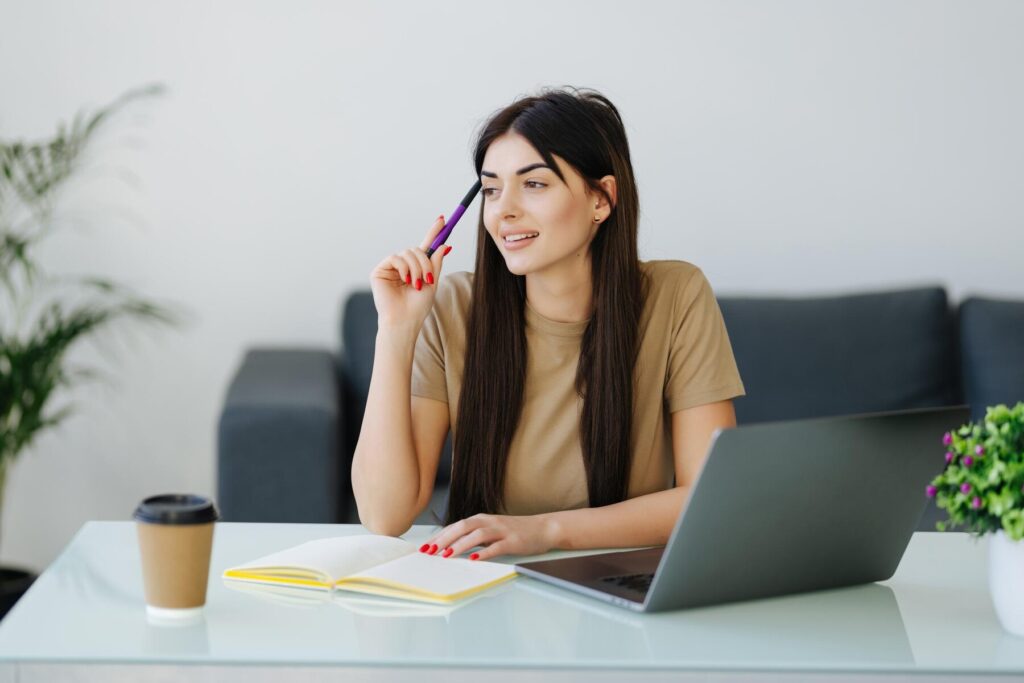 A young woman sitting at a modern desk with a laptop, looking thoughtful as she holds a pen to her chin, with a notebook and coffee cup beside her.