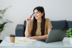 A young woman sitting at a modern desk with a laptop, looking thoughtful as she holds a pen to her chin, with a notebook and coffee cup beside her.