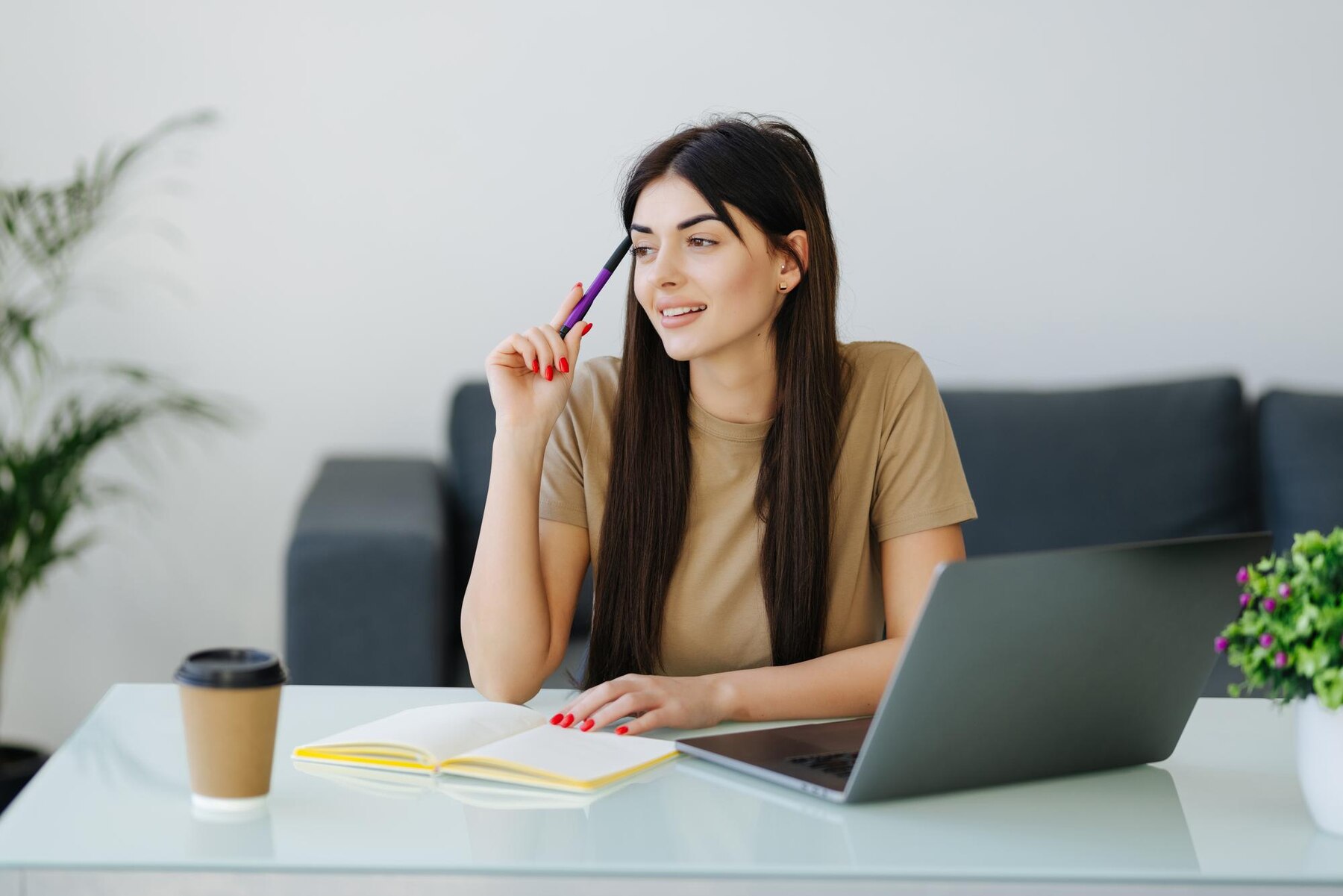 A young woman sitting at a modern desk with a laptop, looking thoughtful as she holds a pen to her chin, with a notebook and coffee cup beside her.