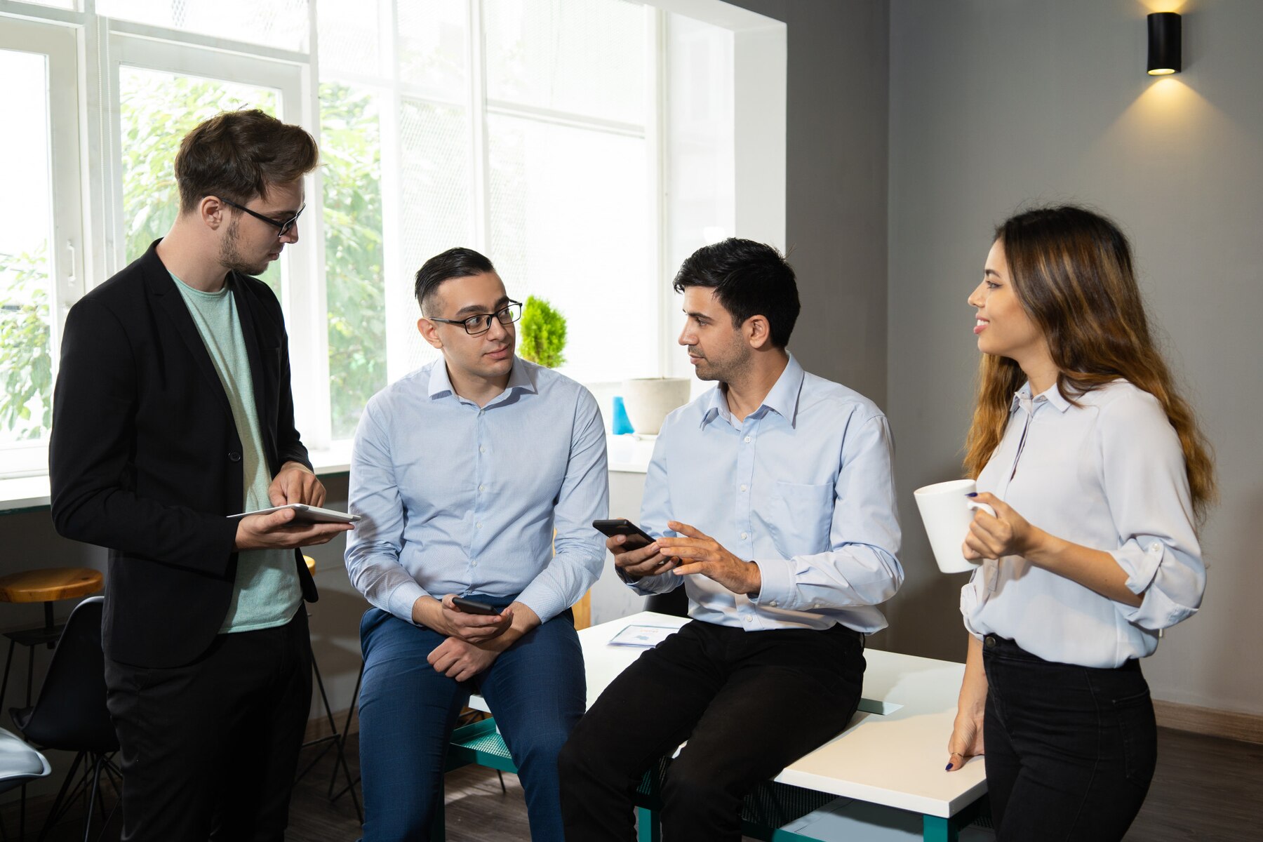 A group of four individuals in casual attire engaged in conversation, sitting on a modern table in a bright, contemporary room.