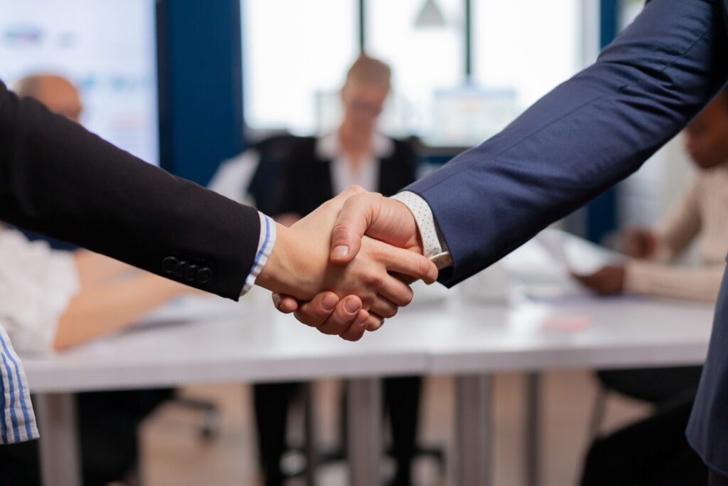 Two hands shake in a business meeting, symbolizing agreement or partnership, with people seated at a table in the background.