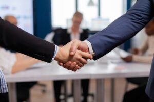 Two hands shake in a business meeting, symbolizing agreement or partnership, with people seated at a table in the background.