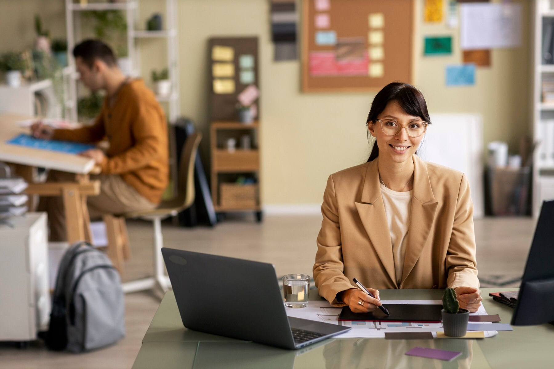 A professional woman in a beige blazer smiles while working at a laptop in a modern office environment, with coworkers visible in the background.