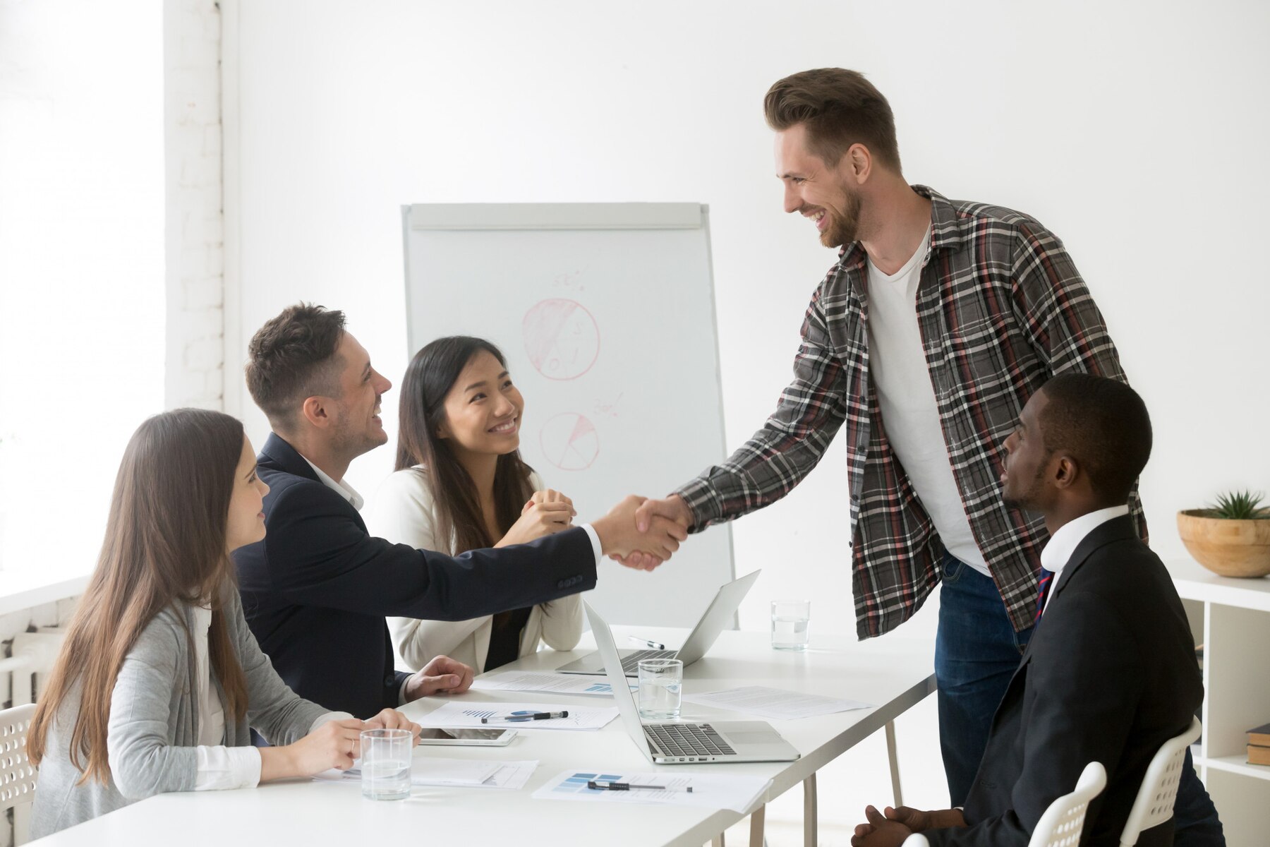 A professional meeting in a bright office, featuring a handshake between two individuals while others observe at the table.