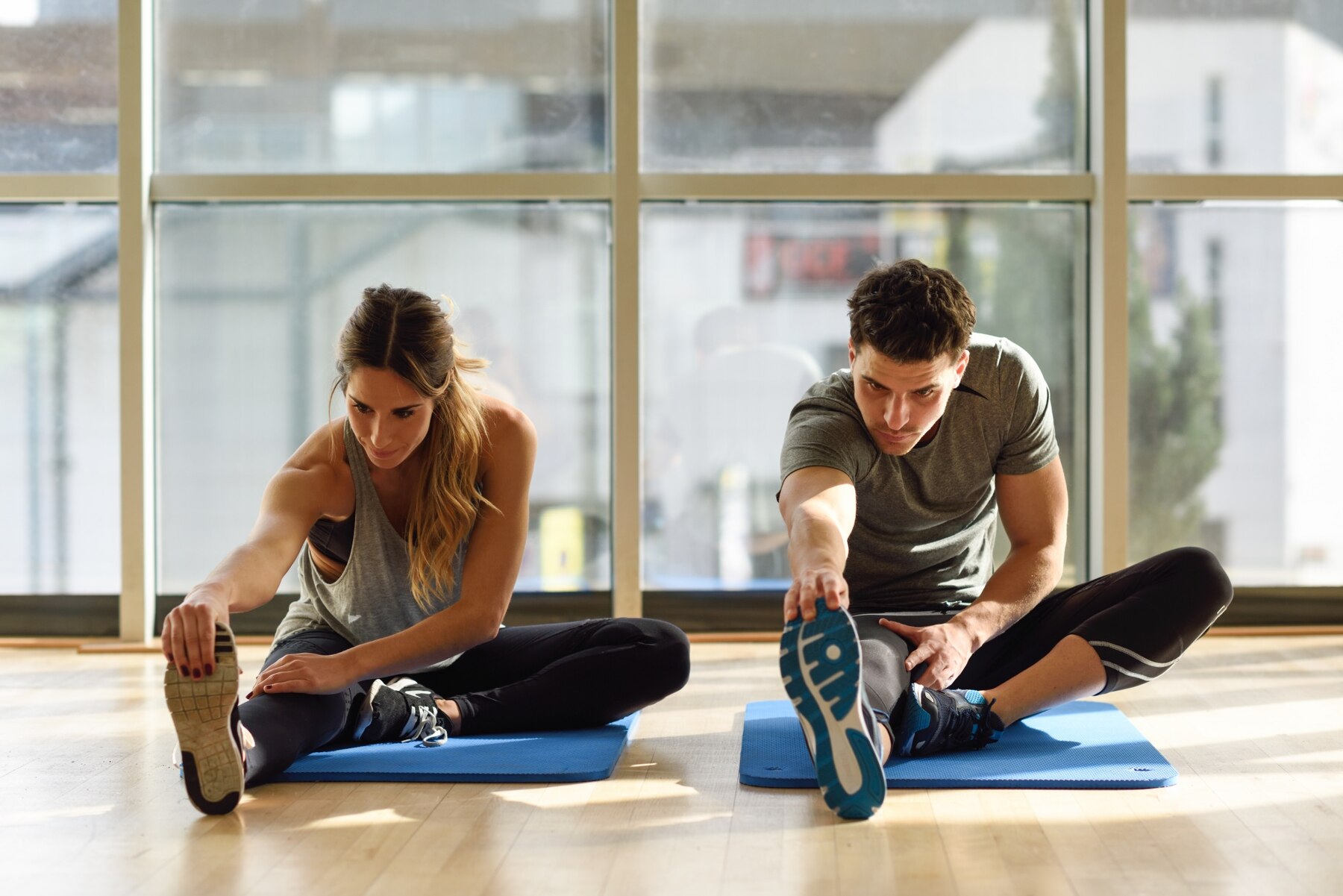 A man and a woman stretching on blue mats in a sunny room, performing seated leg stretches while facing large windows.
