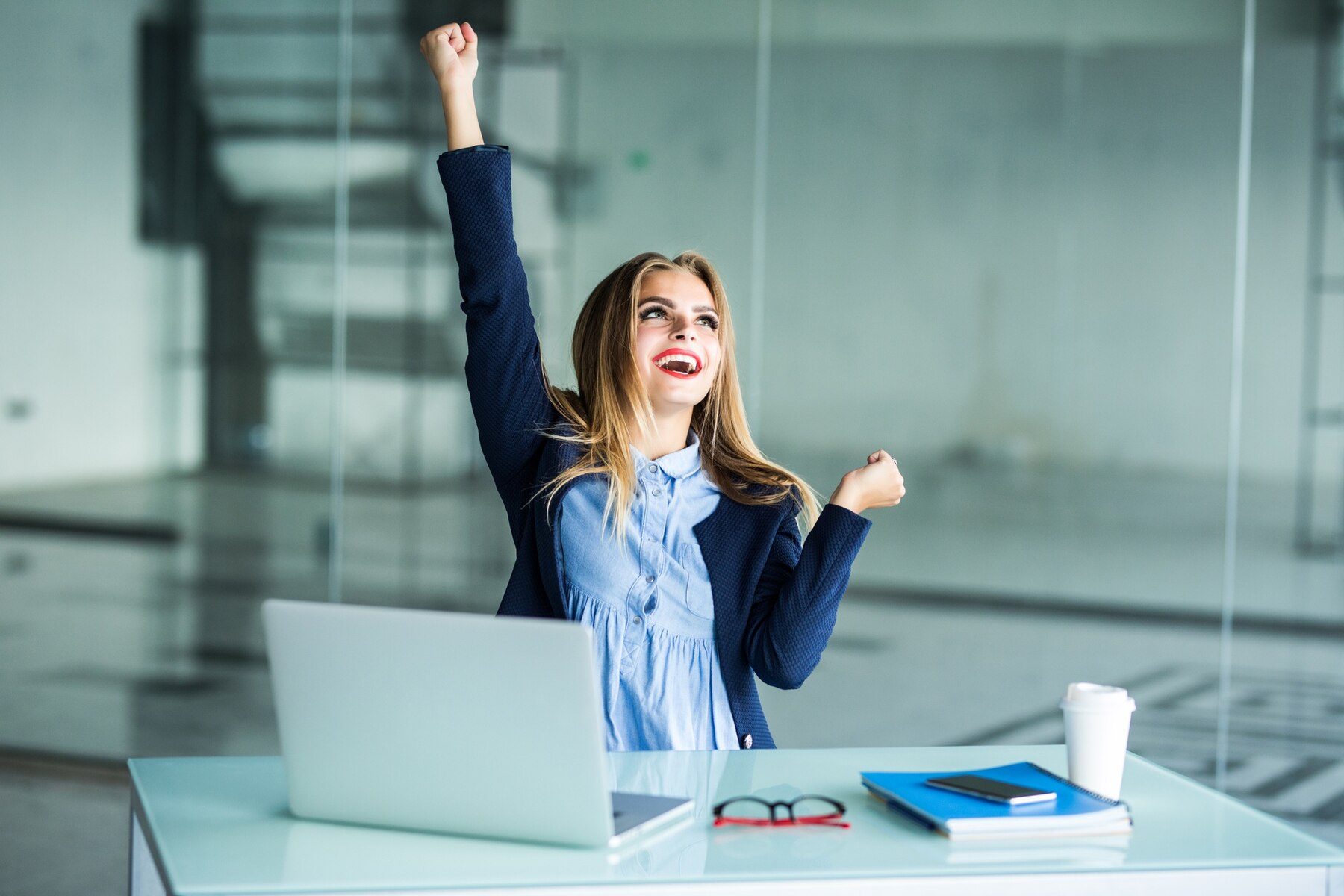 A joyful businesswoman in a modern office, celebrating with arms raised, seated at a desk with a laptop, coffee cup, glasses, and a notebook.