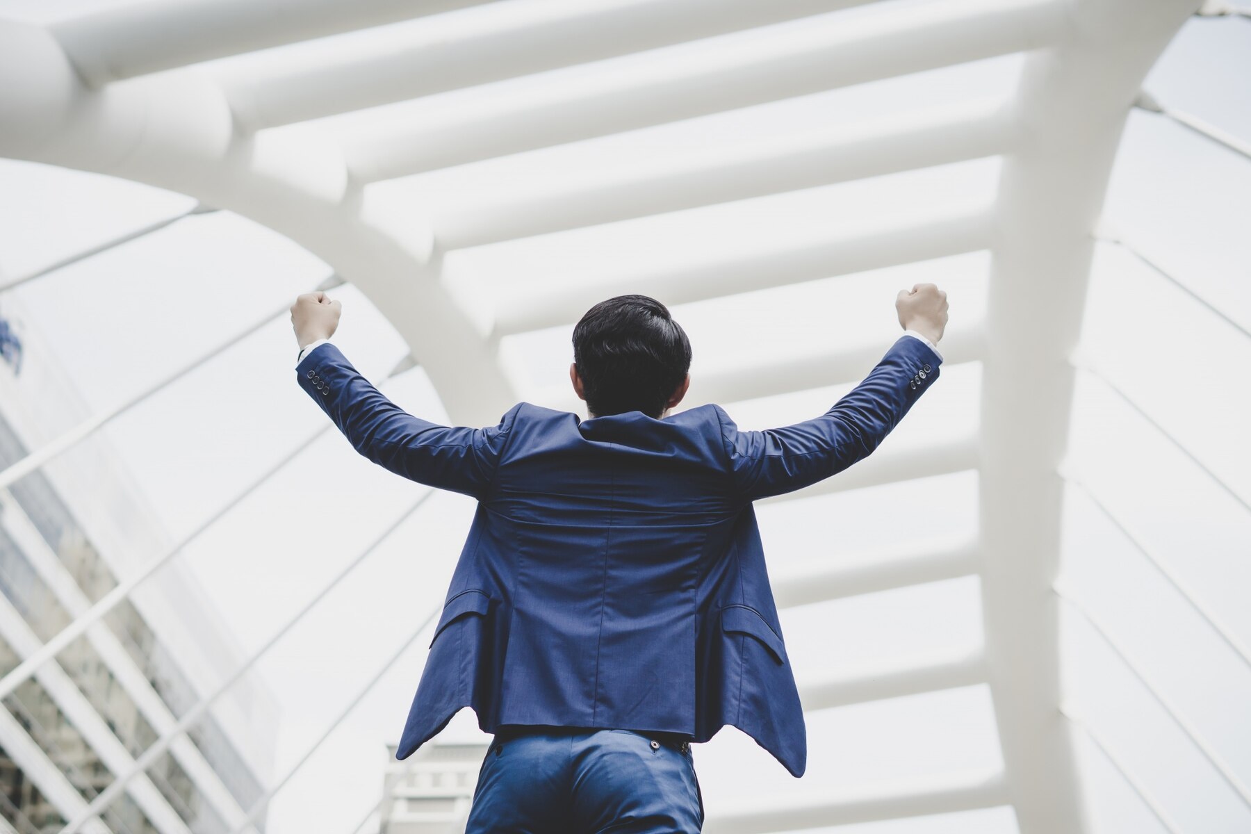 A man in a blue suit with his back to the camera, arms raised in a victorious gesture under a white, modern architectural structure.