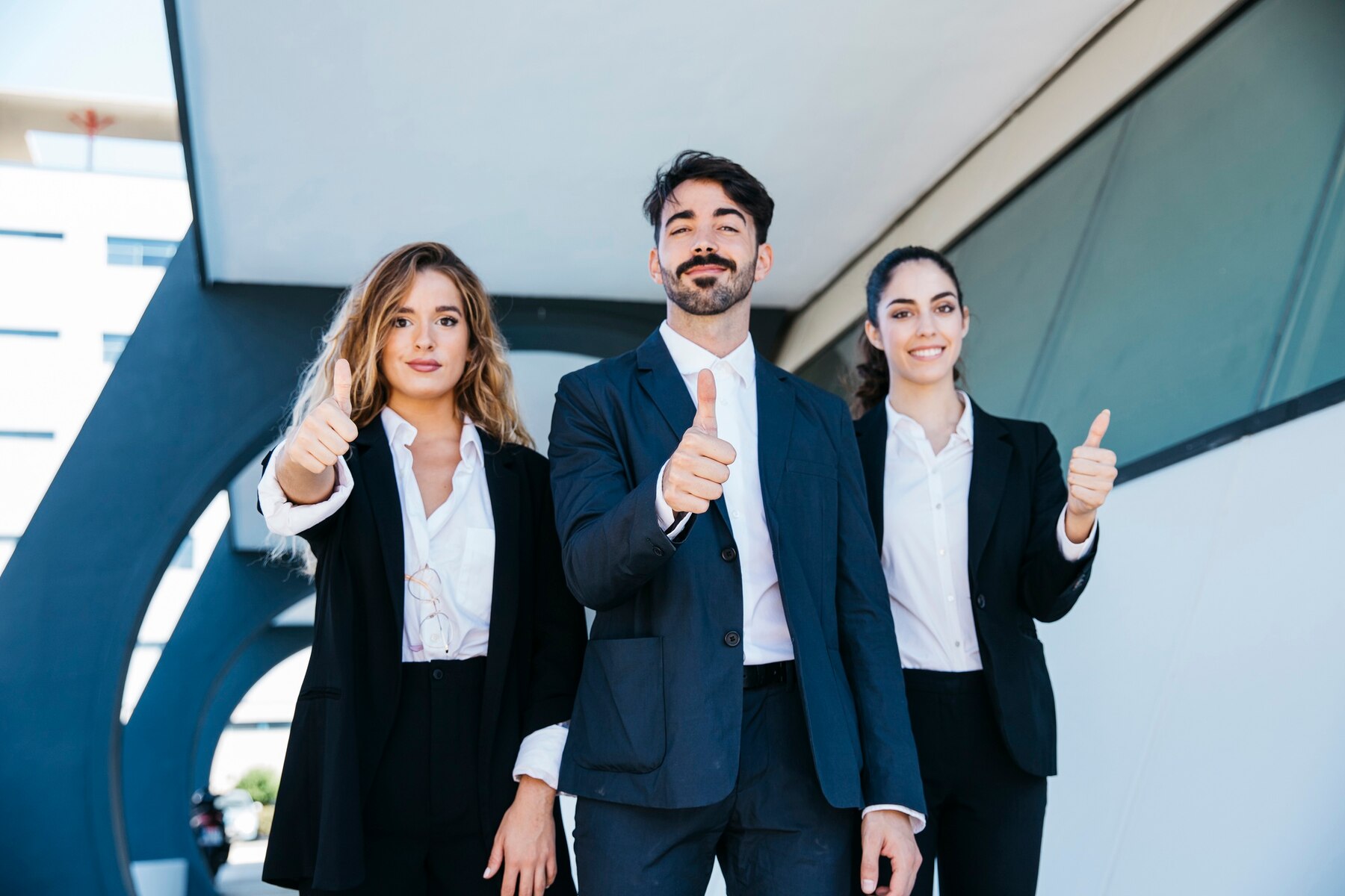 Three young professionals in business suits giving thumbs up in front of a modern building.