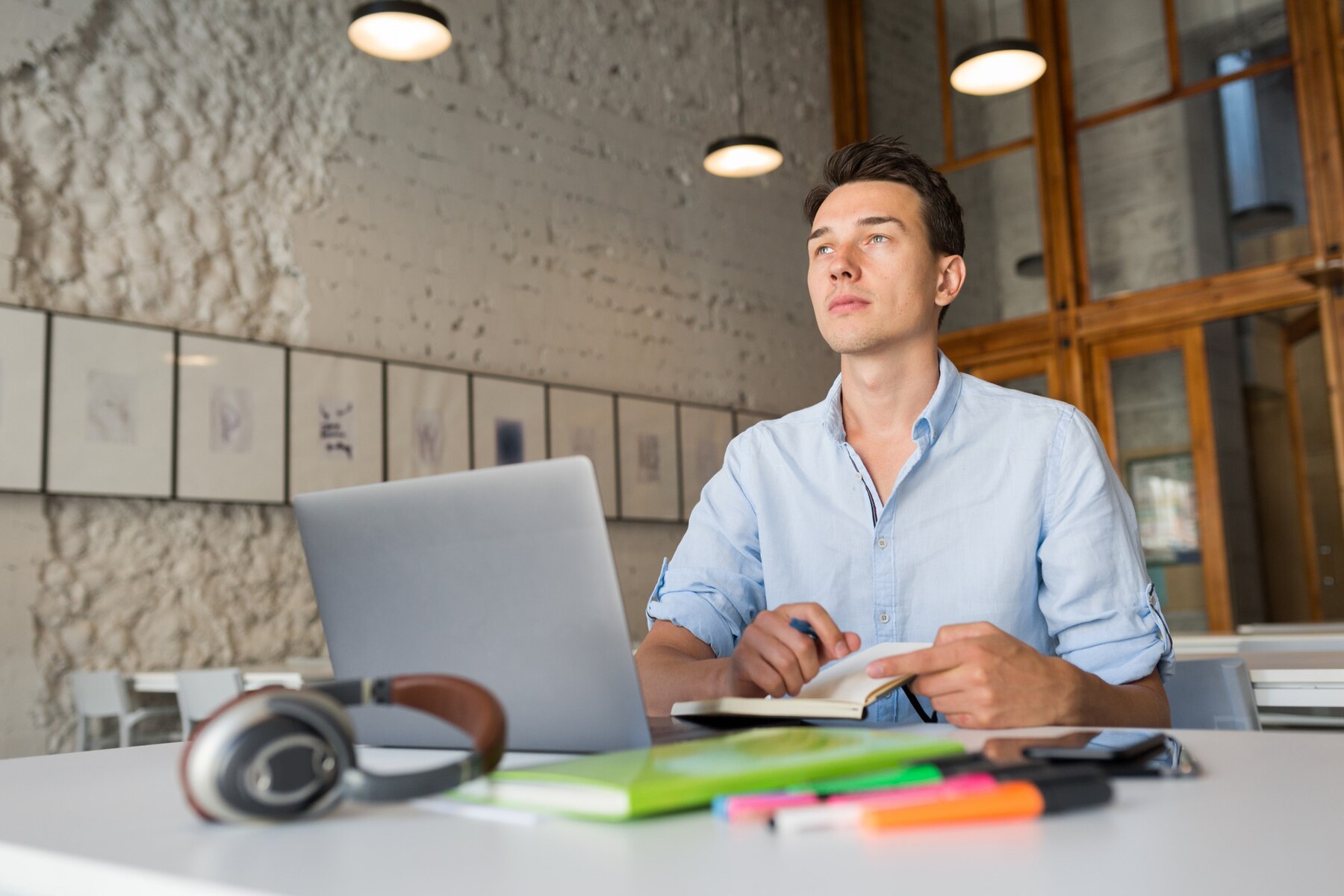 A young man in a light blue shirt sits at a desk in a modern office, looking thoughtfully upwards, with a laptop open in front of him and headphones beside a notebook on the desk.