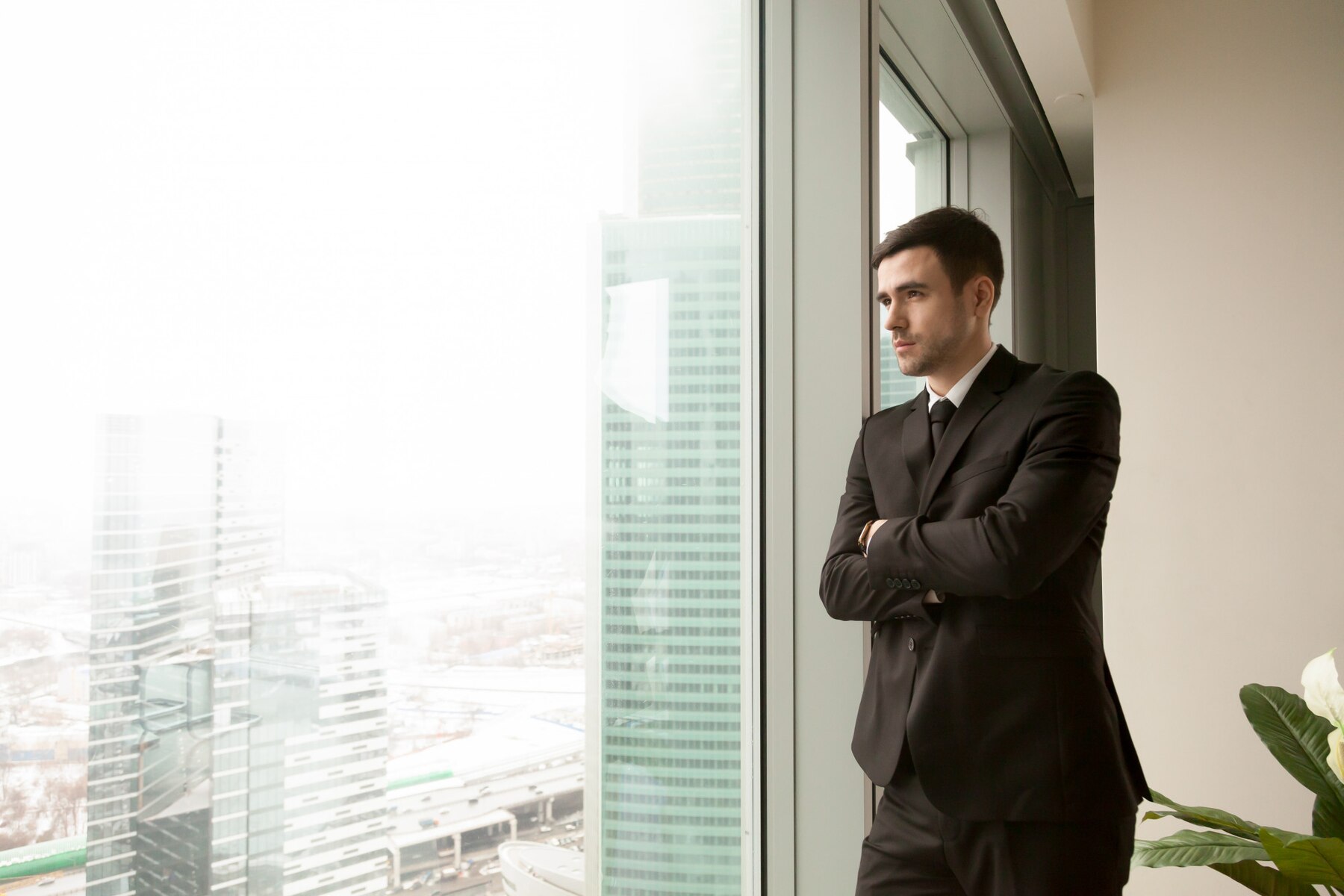 A businessman in a suit stands pensively by a large window overlooking a foggy cityscape.