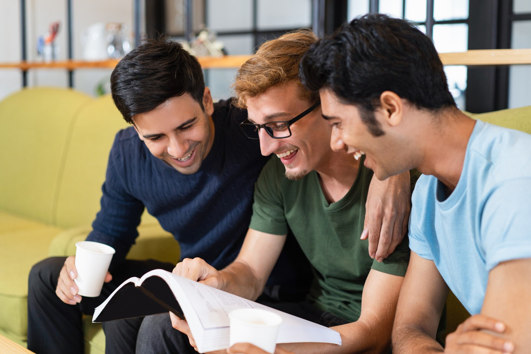 Three young men laughing and looking at a book together on a sofa in a casual, cozy setting.