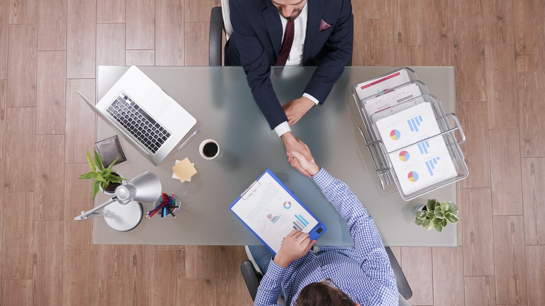 Two business professionals shaking hands over a glass table, with a laptop, coffee, and documents visible in a modern office setting.