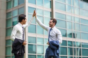 Two businessmen high-fiving in front of a glass office building, expressing joy and collaboration. They are both dressed in formal attire, with one carrying a suit jacket and the other a briefcase.