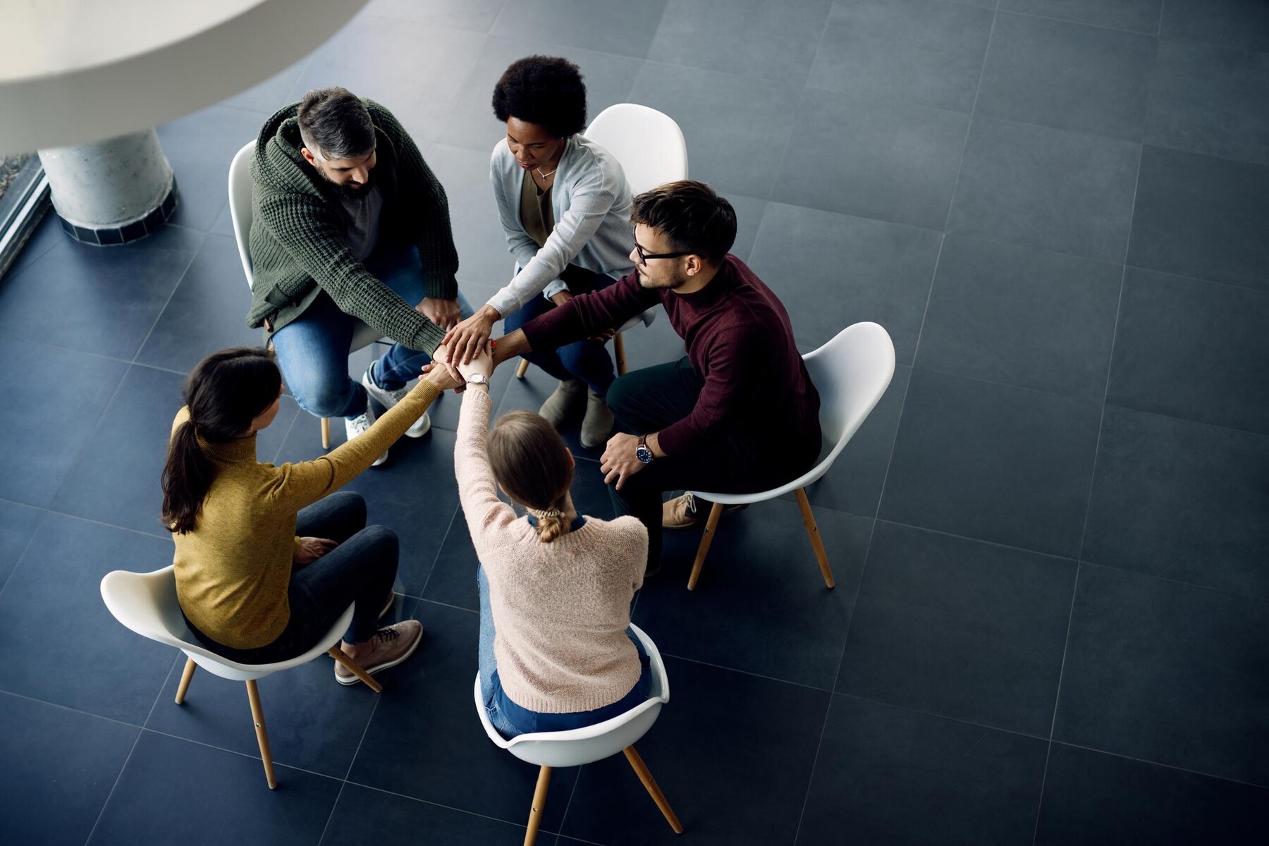 A group of six people seated in a circle, joining hands in a display of teamwork and support in a modern indoor setting.