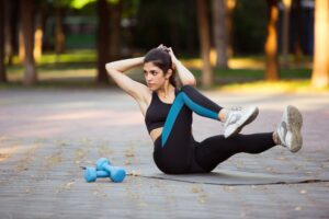 A woman doing a stretching exercise on a paved park path, with a pair of blue dumbbells lying beside her.
