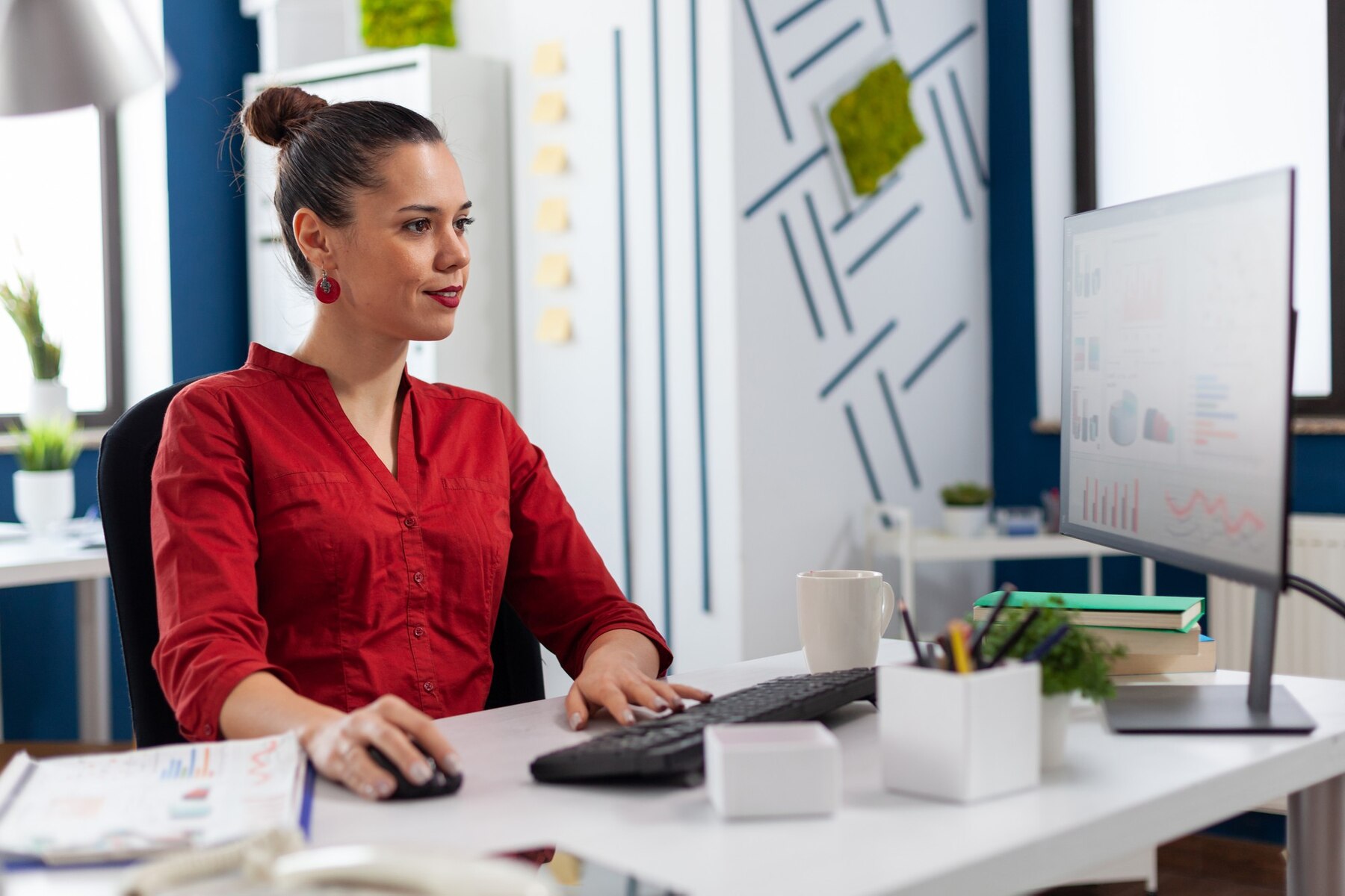 A person in a red shirt works at a desk with a computer, a cup, and green plants in a modern office environment.