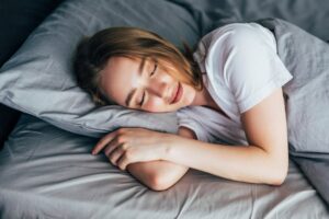 A young woman peacefully sleeping in a grey bed, with her head resting on a pillow and a faint smile on her lips.