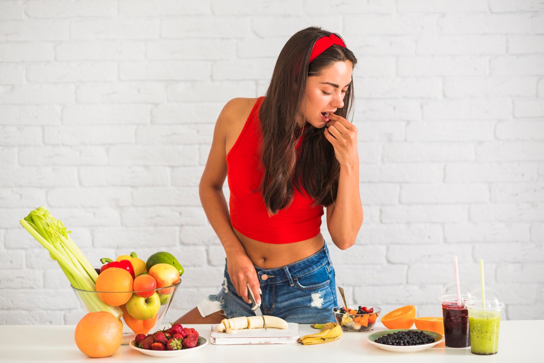 Young woman in a red top and denim shorts eating fresh fruit in a kitchen with a white brick wall background, surrounded by various fruits and smoothies.