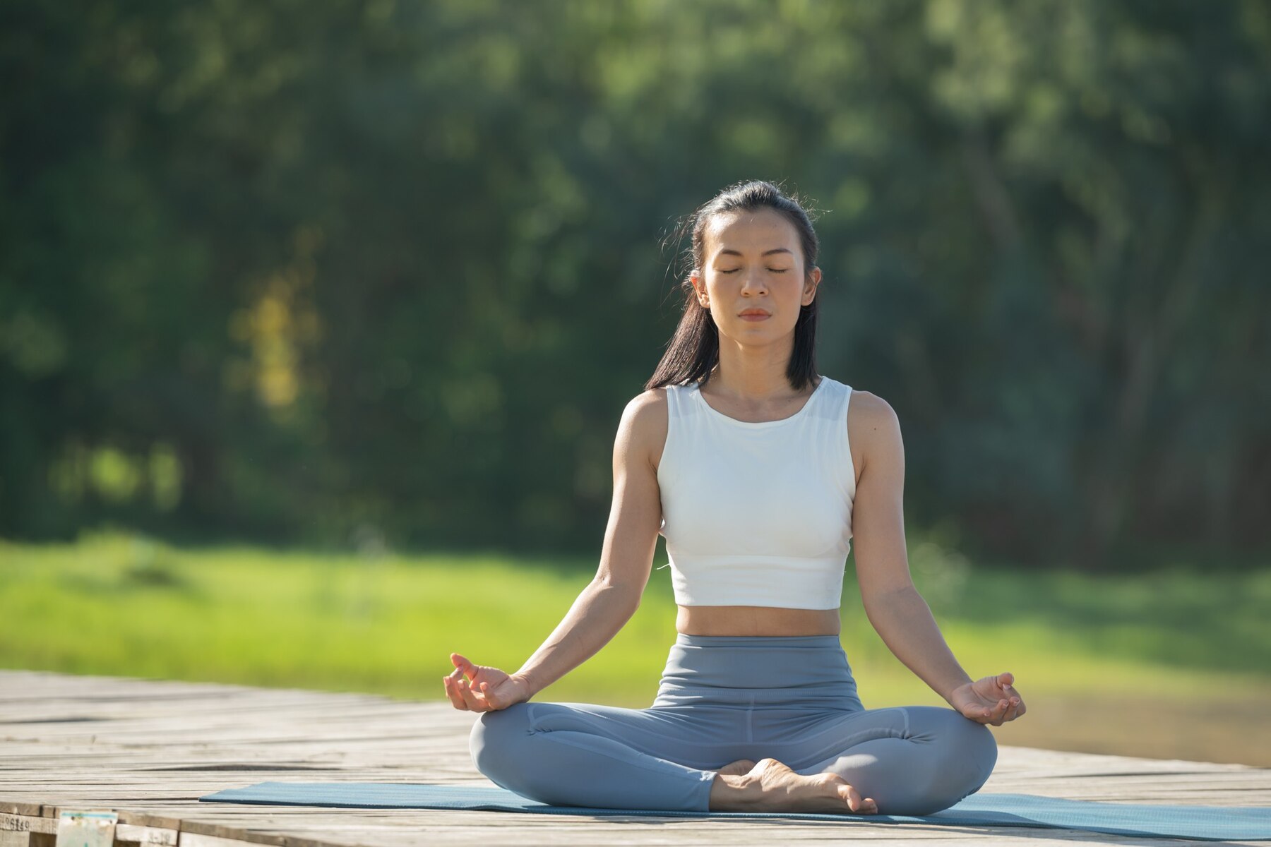 A young woman meditating in a lotus position on a yoga mat outdoors, surrounded by lush greenery, with her eyes closed and a calm expression.