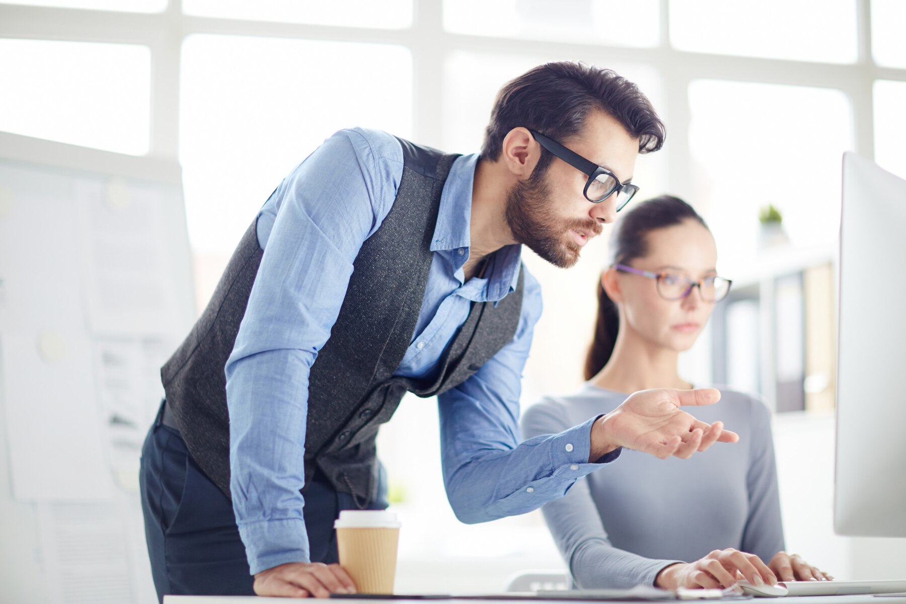 A man in a blue shirt and vest gestures towards a computer screen while discussing with a woman in a grey sweater.