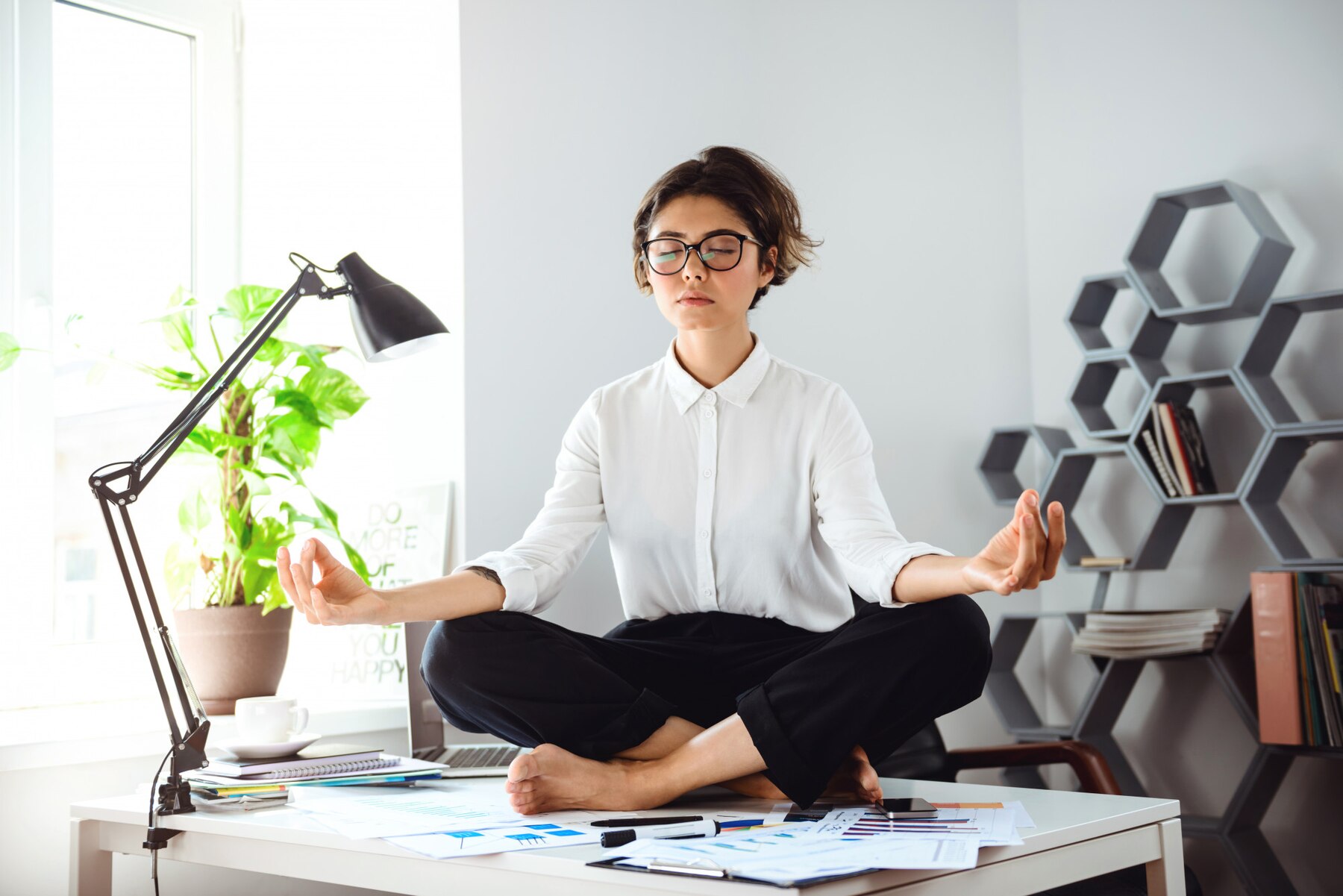 A young woman in business attire sitting cross-legged on a desk, meditating with her eyes closed, surrounded by documents and a laptop, in a well-lit modern office.