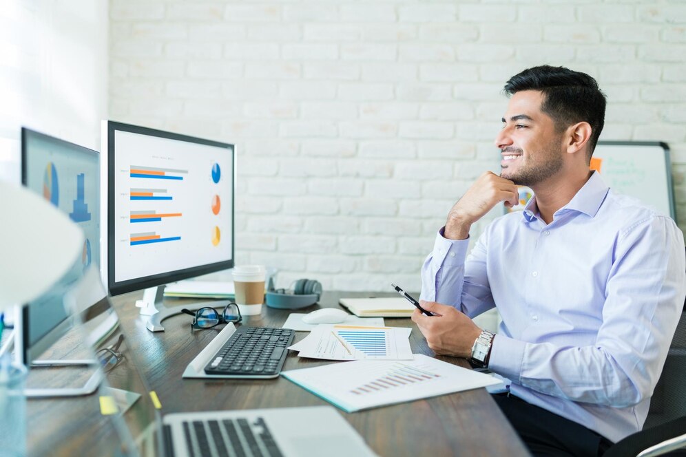 A man in a light blue shirt sits at a desk, analyzing charts on dual monitors amidst office supplies and a coffee cup.
