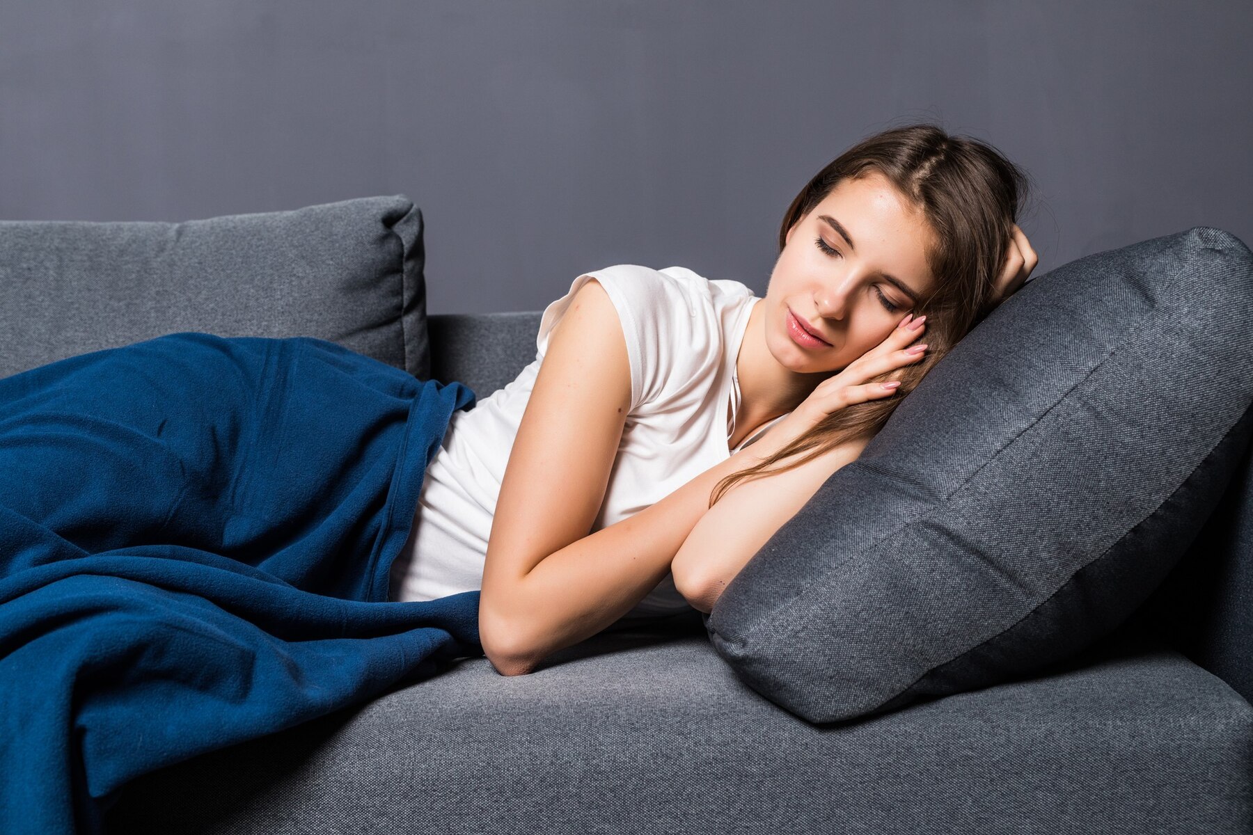 Young woman sleeping peacefully on a grey sofa, covered with a blue blanket.