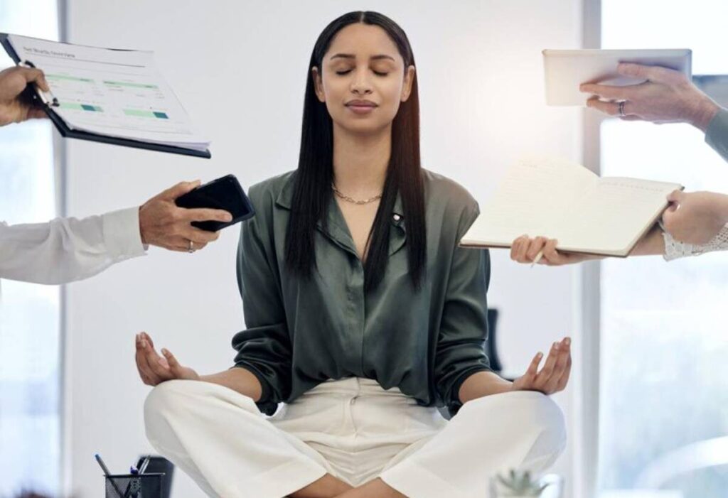 A person meditates in an office setting, surrounded by hands offering documents, tablets, and notebooks, emphasizing a busy work environment.
