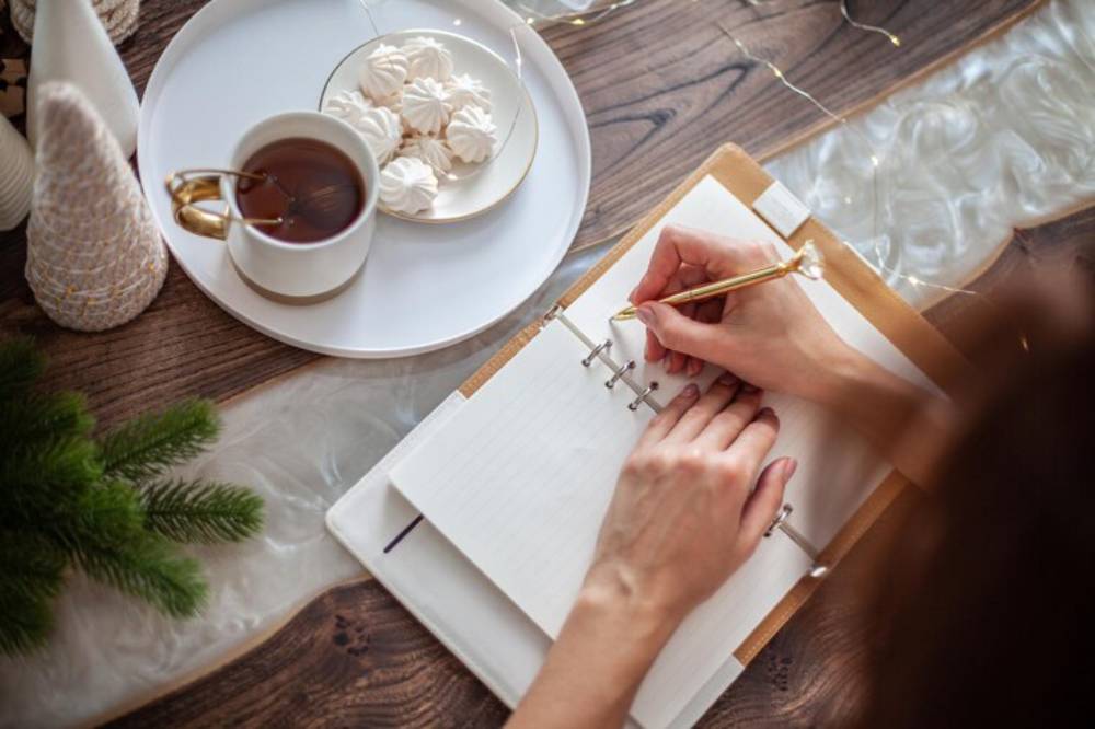 A person writes in a planner with a gold pen, alongside a cup of tea and a plate of meringues on a wooden table adorned with greenery.