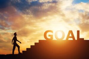 Silhouette of a person climbing stairs towards a large "GOAL" sign against a sunset sky, symbolizing achievement and aspiration.