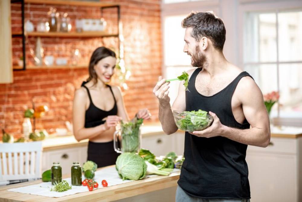 A man in a black tank top holds a bowl of salad while a woman prepares vegetables in a cozy kitchen with brick walls.