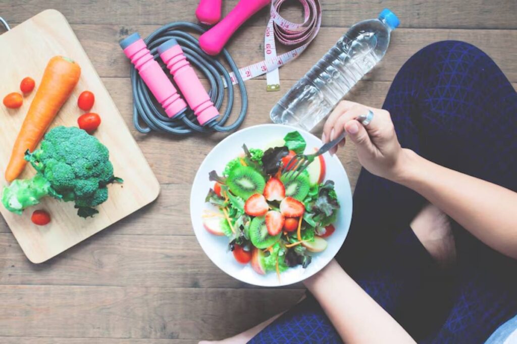 A person sitting on the floor enjoys a colorful salad, surrounded by fresh vegetables, a water bottle, and fitness equipment.