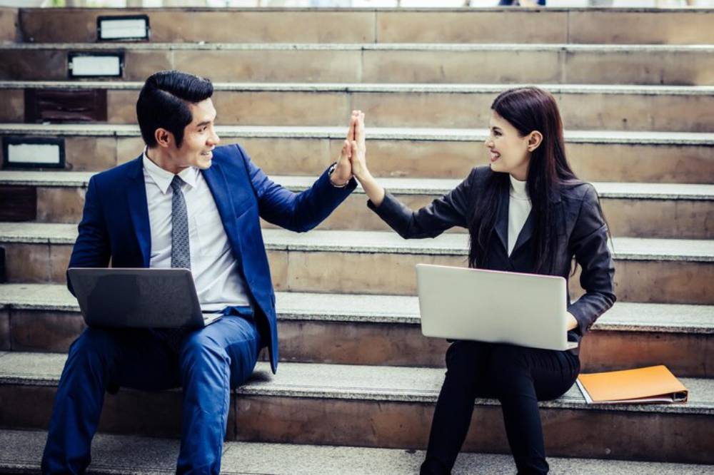 A man in a suit and a woman in a blazer sit on stairs, engaging in a high-five gesture while working on laptops.