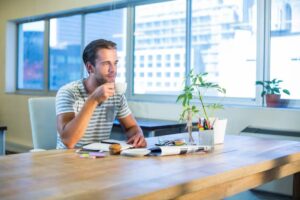 A person sits at a wooden table, sipping coffee, surrounded by office supplies and a small plant, with large windows in the background.