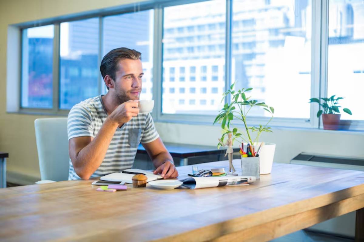 A person sits at a wooden table, sipping coffee, surrounded by office supplies and a small plant, with large windows in the background.