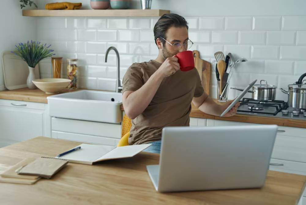 A person sits at a wooden kitchen table, sipping from a red mug and holding a tablet, with a laptop and kitchen items nearby.