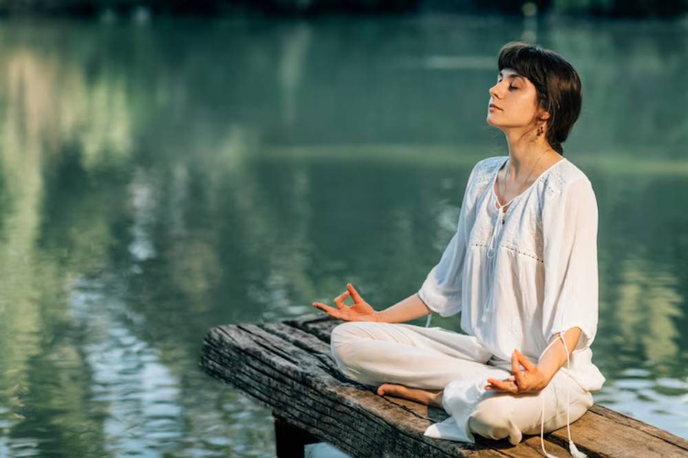 A person dressed in white meditating peacefully on a wooden dock by a serene lake, surrounded by lush green nature.