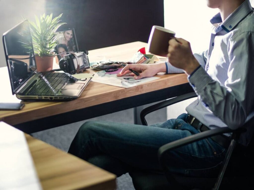 A person sits at a desk, holding a mug, surrounded by a laptop, camera, plant, and scattered papers, in a well-lit workspace.