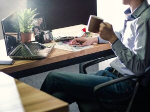A person sits at a desk, holding a mug, surrounded by a laptop, camera, plant, and scattered papers, in a well-lit workspace.