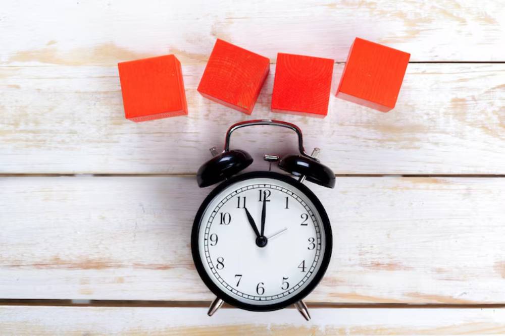 A black alarm clock with a white face sits on a wooden surface, accompanied by four bright red blocks.